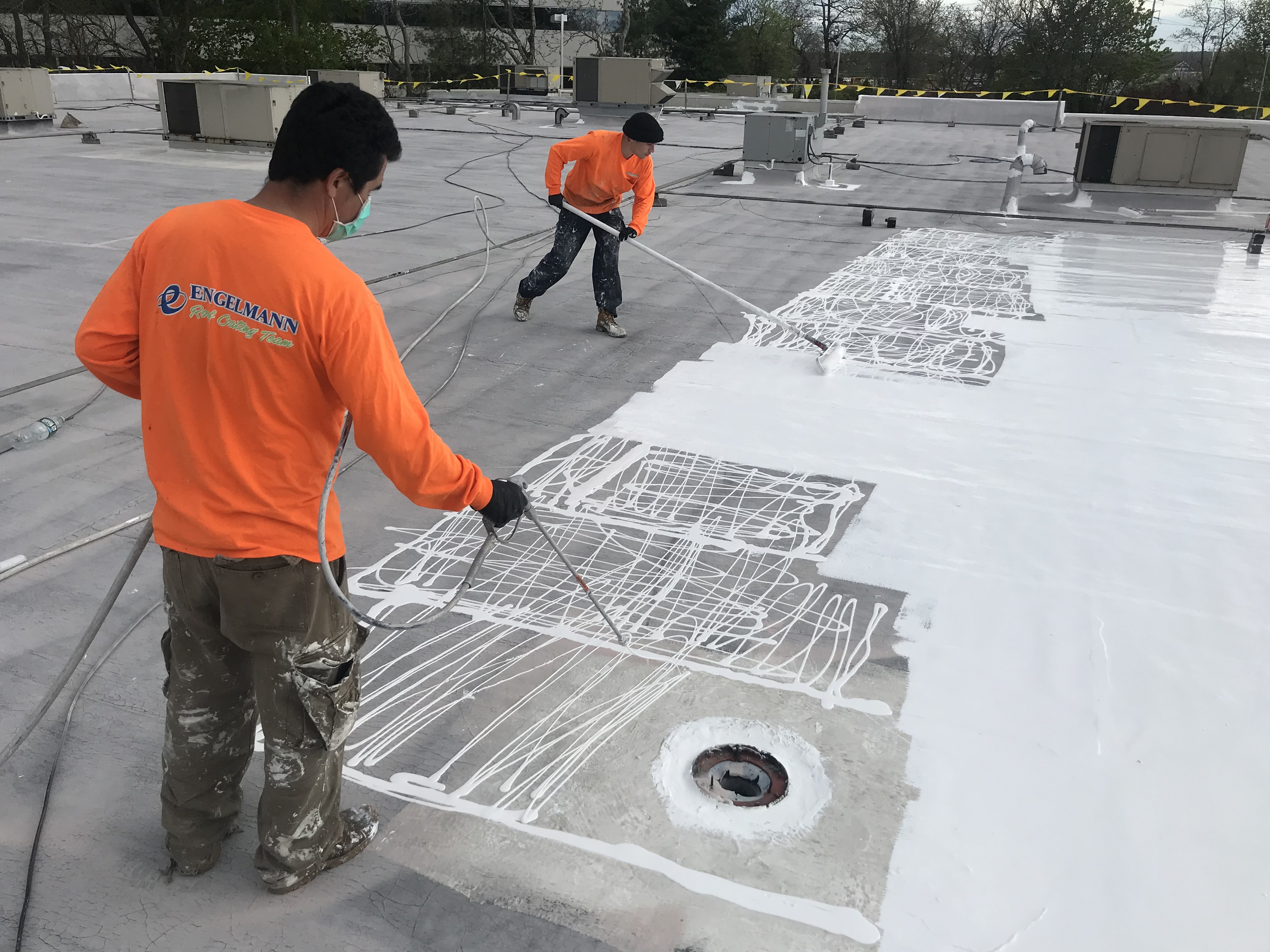 Engelmann Construction worker spraying a white liquid coating on a commercial roof while another spreads it out with a roller